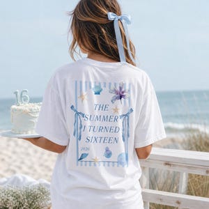 Könnte beinhalten: Ein weißes T-Shirt mit einer blau gestreiften rechteckigen Grafik auf der Rückseite mit dem Text "THE SUMMER I TURNED SIXTEEN 2026". Das Design enthält Aquarellillustrationen einer violetten Blume, Muscheln, Seesternen und blauen Schleifen.