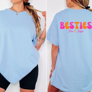 May include: Light blue t-shirt with the word "BESTIES" in pink and orange bubble letters on the back, with the names "Pia & Jessie" below. The shirt is a casual style.