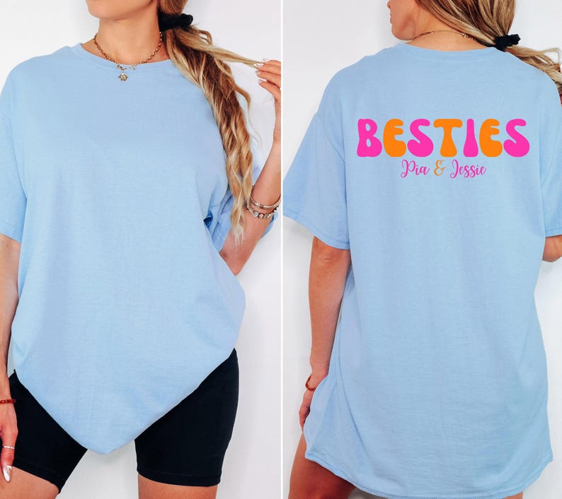 May include: Light blue t-shirt with the word "BESTIES" in pink and orange bubble letters on the back, with the names "Pia & Jessie" below. The shirt is a casual style.