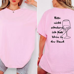 May include: A pale pink t-shirt with short sleeves. The back of the shirt features black text and a line drawing of a hand holding a wine glass. The text reads "Bitte nicht schubsen, ich hab Wein in der Hand."