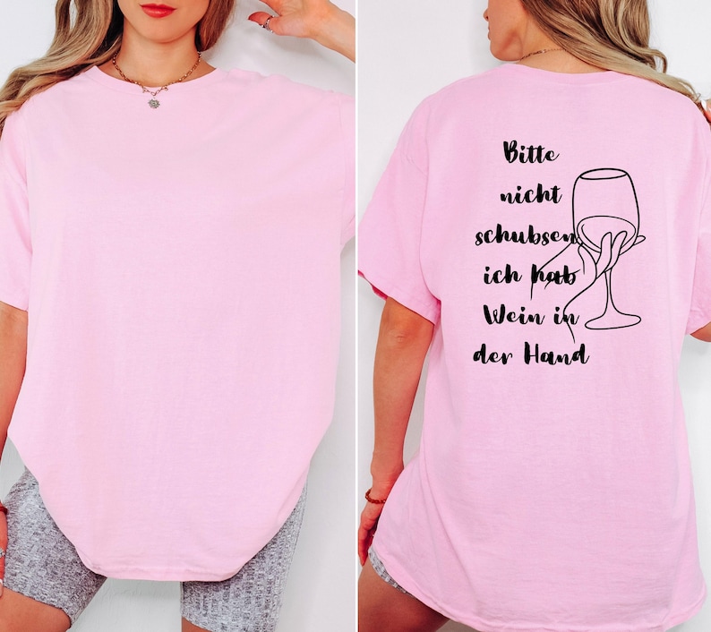May include: A pale pink t-shirt with short sleeves. The back of the shirt features black text and a line drawing of a hand holding a wine glass. The text reads "Bitte nicht schubsen, ich hab Wein in der Hand."