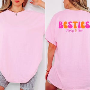 May include: A light pink t-shirt with the word "BESTIES" in orange and pink lettering on the back. The names "Franzi & Thea" are printed below the word "BESTIES". The front of the shirt is plain.