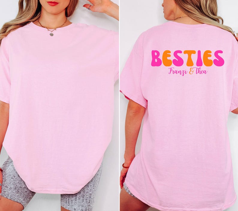 May include: A light pink t-shirt with the word "BESTIES" in orange and pink lettering on the back. The names "Franzi & Thea" are printed below the word "BESTIES". The front of the shirt is plain.