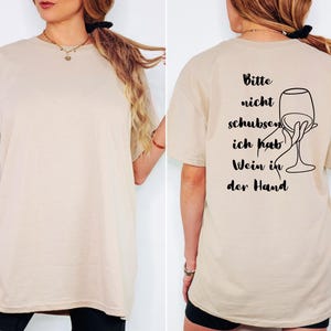 May include: Beige t-shirt featuring the German phrase "Bitte nicht schubsen, ich hab Wein in der Hand" and a wine glass illustration. The shirt is a neutral colour and has a loose fit.
