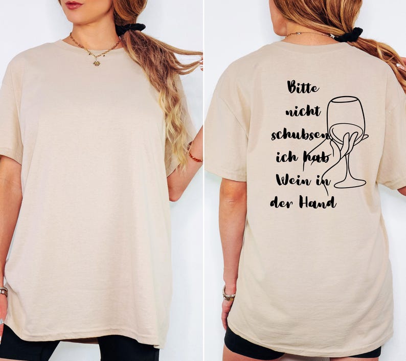 May include: Beige t-shirt featuring the German phrase "Bitte nicht schubsen, ich hab Wein in der Hand" and a wine glass illustration. The shirt is a neutral colour and has a loose fit.