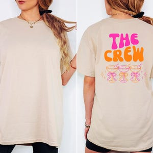 May include: Beige t-shirt with the text "THE CREW" in pink and orange on the back, along with three cocktail glasses. The front of the shirt is plain. The shirt is a casual, oversized fit.
