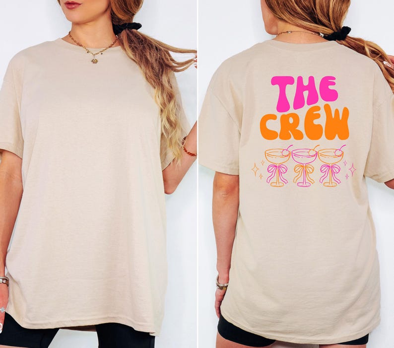May include: Beige t-shirt with the text "THE CREW" in pink and orange on the back, along with three cocktail glasses. The front of the shirt is plain. The shirt is a casual, oversized fit.