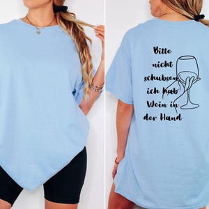 May include: Pale blue t-shirt featuring the German phrase "Bitte nicht schubsen, ich hab Wein in der Hand" and a wine glass illustration on the back. The front is plain. The shirt is short-sleeved and of a relaxed fit.