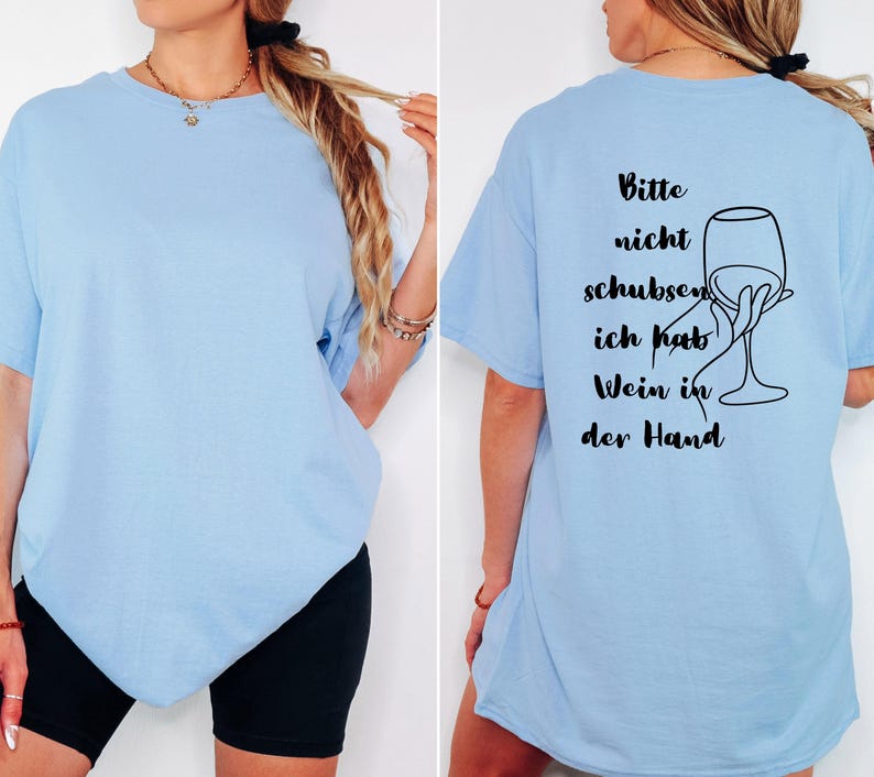 May include: Pale blue t-shirt featuring the German phrase "Bitte nicht schubsen, ich hab Wein in der Hand" and a wine glass illustration on the back. The front is plain. The shirt is short-sleeved and of a relaxed fit.