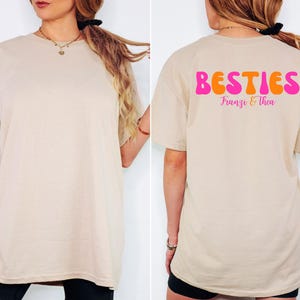 May include: Beige t-shirt with the word "BESTIES" in orange and pink bubble letters on the back, with the names "Franzi & Thea" below. The front of the shirt is plain.