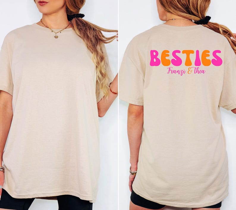 May include: Beige t-shirt with the word "BESTIES" in orange and pink bubble letters on the back, with the names "Franzi & Thea" below. The front of the shirt is plain.