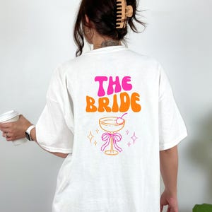May include: White t-shirt with the text "THE BRIDE" in pink and orange retro font. Below the text is a cocktail glass with a cherry, a pink bow, and star accents. The shirt is a relaxed fit.