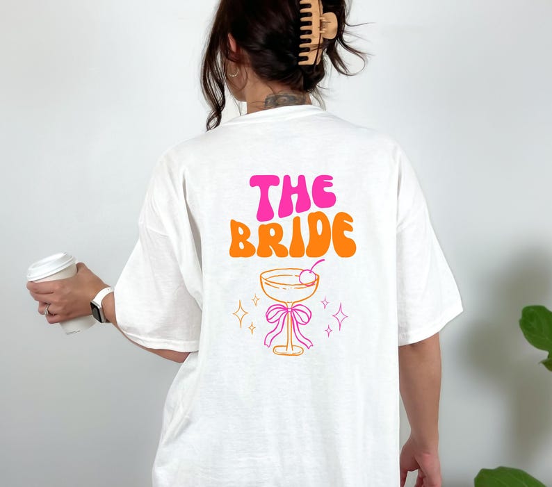 May include: White t-shirt with the text "THE BRIDE" in pink and orange retro font. Below the text is a cocktail glass with a cherry, a pink bow, and star accents. The shirt is a relaxed fit.