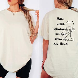 May include: A cream-coloured t-shirt featuring the German phrase "Bitte nicht schubsen, ich hab Wein in der Hand" and a line drawing of a hand holding a wine glass. The shirt has a relaxed, oversized fit.