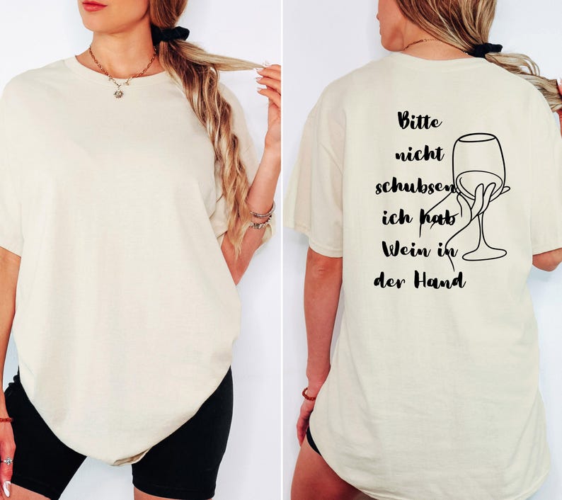 May include: A cream-coloured t-shirt featuring the German phrase "Bitte nicht schubsen, ich hab Wein in der Hand" and a line drawing of a hand holding a wine glass. The shirt has a relaxed, oversized fit.