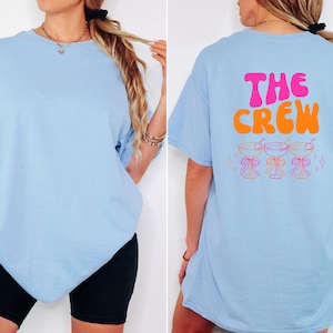 May include: Light blue t-shirt with short sleeves. The front of the shirt is plain. The back of the shirt has the words "THE CREW" in pink and orange, with three cocktail glasses below. The shirt is a casual style.