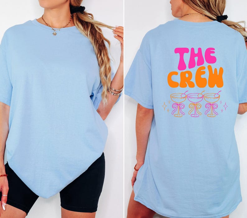 May include: Light blue t-shirt with short sleeves. The front of the shirt is plain. The back of the shirt has the words "THE CREW" in pink and orange, with three cocktail glasses below. The shirt is a casual style.