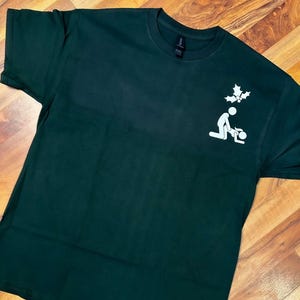 May include: A dark green t-shirt with a white graphic of a stick figure kneeling with a holly sprig above their head.