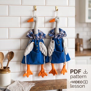 Op de afbeelding: Twee hangende stoffen ganzenversieringen met grijze koppen, oranje snavels en voeten, en lichamen van blauwe corduroy. Elke gans draagt een blauwe geruite ruchekraag, heeft witte stoffen vleugels en drie kleine houten knoopjes op de borst.