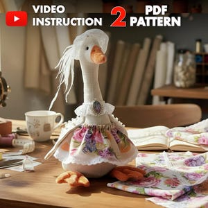May include: A handmade goose doll with a white head, neck, and body, wearing a floral dress and a white hat. The goose has an orange beak and feet. The image includes the text "VIDEO INSTRUCTION 2 PDF PATTERN".