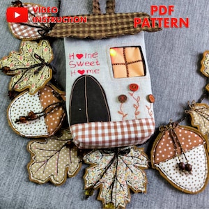 Può includere: Una casa di stoffa fatta a mano con la scritta "Home Sweet Home" e una finestra. La casa è circondata da foglie e ghiande decorative. L'immagine include anche il testo "Video Instruction" e "PDF Pattern".