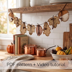 May include: A fabric garland featuring alternating autumn leaf and acorn shapes in various brown, tan, and cream patterns, suspended by a dark string. The leaves and acorns are stitched with visible edges and small wooden bead accents throughout the design.