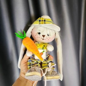 Pode incluir: Boneca de coelho de pelúcia feita à mão com orelhas compridas, usando um chapéu e macacão xadrez amarelo. O coelho segura uma grande cenoura laranja. A boneca tem um rosto branco com sardas e sapatos de feltro cinza.