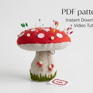 Könnte beinhalten: Ein pilzförmiges Nadelkissen mit rotem Hut und weißen Punkten, besetzt mit bunten Nadeln. Der Stiel ist beige, mit grünem Sockel und dekorativen Perlen. Der Text "PDF pattern" ist sichtbar.