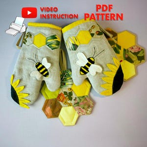 May include: A pair of gray oven mitts with yellow trim, featuring bee and sunflower appliqués. The mitts are part of a set that includes a matching honeycomb-shaped hot pad. The words "VIDEO INSTRUCTION PATTERN PDF" are printed at the top.