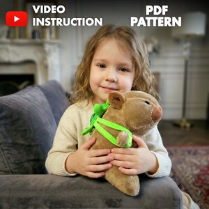 Peut inclure: Un jouet en peluche en forme de cochon marron avec des sangles vertes et un nœud vert est tenu par un jeune enfant. L'image comprend le texte "VIDEO INSTRUCTION" et "PDF PATTERN". L'enfant est assis sur un canapé gris.