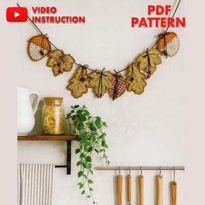 Könnte beinhalten: Eine handgefertigte Girlande zum Thema Herbst mit Eichel- und Blattformen in Braun-, Beige- und Orangetönen, auf einer schwarzen Schnur aufgereiht. Die Girlande wird vor einer weißen Wand mit dem Text "VIDEO INSTRUCTION" und "PDF PATTERN" angezeigt.