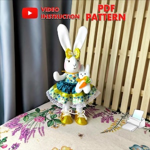 May include: A white fabric bunny doll wearing a green and yellow dress with a yellow bow. The bunny is holding a smaller white fabric bunny doll with a carrot. The background is a wooden slat wall with a printer icon and the text "PDF PATTERN" and "VIDEO INSTRUCTION".