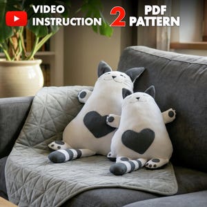 Op de afbeelding: Twee witte pluche kattenspeeltjes met donkergrijze oren, staarten en hartvormige vlekken op hun borst. De katten hebben geborduurde gezichten met gesloten ogen en kleine glimlachjes. De tekst bovenaan luidt VIDEO INSTRUCTION en 2 PDF PATTERN.