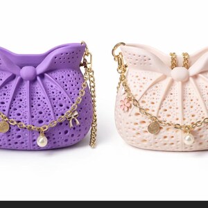 Può includere: Due borse decorative, una lavanda e una rosa pallido, ciascuna con una tracolla a catena dorata e ciondoli decorativi. Le borse hanno un design traforato unico e un dettaglio a fiocco.