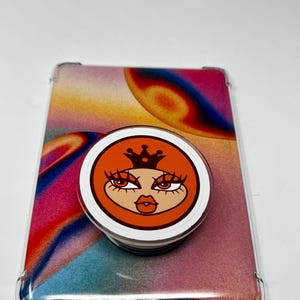 May include: A clear phone case with a colorful abstract design and a pop-up phone grip. The pop-up grip features a cartoon face with orange hair, a crown, and long eyelashes. The case and grip are on a white surface.