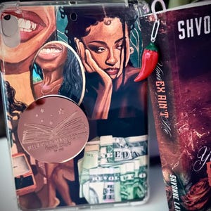 May include: A clear tablet case featuring a collage of illustrations of people, a stack of money, and a circular rose gold pop-out grip with the text Melanin & Manicures Book Club est 2014. A paperclip bookmark with a red chili charm is attached.