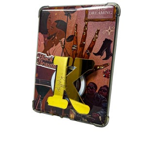 May include: A rectangular phone case featuring a collage of illustrations including a couple, hands with gold rings, and the text "Black Beauty" and "Never Stop Dreaming." A large, reflective gold-colored letter "K" is attached to the center.