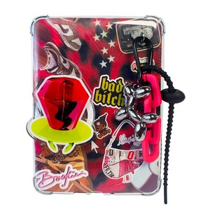May include: A clear protective tablet case featuring a collage of stickers including a red ring pop, a sneaker, a pager, and text reading bad bitch and Bookie. A black carabiner with a silver balloon dog charm and a pink plastic chain is attached.