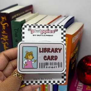 Puede incluir: Una pegatina de tarjeta de biblioteca rosa y blanca con un personaje de dibujos animados del programa "Arthur". La pegatina tiene el texto "Library Card" y un código de barras con el número "2101607". La pegatina es de la empresa "poppies" de "Outta Print".