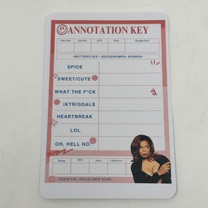 May include: A white card with a red border and a smiling face in the top left corner. The card has the text "Annotation Key" in red letters. The card has a list of terms with icons next to them, including "Sweet/Cute", "What the F*ck", "IKTR/Goals", "Heartbreak", "LOL", and "Oh, Hell No.". The card also has a table with the headings "Rating", "Spice", "Story", and "Characters". The card has a picture of a woman with dark hair and a black top in the bottom right corner.