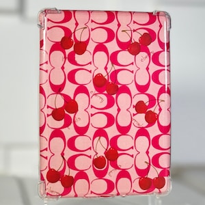 May include: A pink and red phone case with a repeating pattern of the Coach logo and cherries. The cherries are red and have green stems. The text "cherry" is printed on the case.