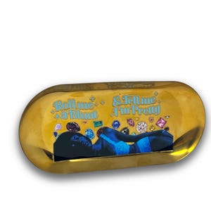 May include: Gold rolling tray with a woman lying down on it. The tray has the text "Roll me a blunt & Tell me I'm Pretty" printed on it. The woman is surrounded by colorful jewels.