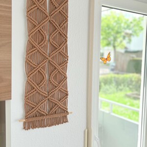 Könnte beinhalten: Ein Wandbehang aus Makramee in hellem Braun mit einem geometrischen Rautenmuster. Das Textilkunstwerk hängt an einem Holzstab vor einer weißen Wand. Dieses dekorative Element verleiht jedem Raum einen Hauch von Bohème-Stil, ca. 70 cm lang.
