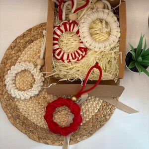 Könnte beinhalten: Eine Sammlung handgefertigter Makramee-Kranz-Ornamente in einer Pappschachtel. Die Kränze sind in verschiedenen Farben, darunter Rot und Creme, und werden auf einer gewebten Matte präsentiert. Die Schachtel ist mit geschreddertem Papier gefüllt.