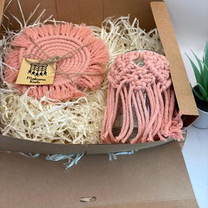 Könnte beinhalten: Ein korallenfarbenes Makramee-Untersetzer- und Gläser-Set in einer Pappschachtel mit Papierfüllung. Der Untersetzer ist rund mit einem Fransenrand. Die Gläserhülle hat eine gewebte Oberseite und lange Fransen. Ein kleines Etikett trägt die Aufschrift "Makrama Knots".