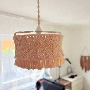 Puede incluir: Lámpara colgante de macramé beige. La pantalla presenta un patrón geométrico de diamantes y un ribete de flecos. La luz cuelga de un cable trenzado y está suspendida de una lámpara de techo blanca. El estilo general es bohemio.