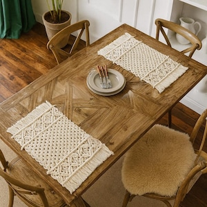 Könnte beinhalten: Ein Holztisch mit zwei cremefarbenen Makramee-Tischsets, jedes mit einem Fransenrand. Der Tisch ist mit Tellern und Besteck gedeckt. Holzstühle umgeben den Tisch, einer mit einem Schaffell-Sitzbezug. Ein Topf mit Pflanzen und ein grüner Vorhang befinden sich im Hintergrund.