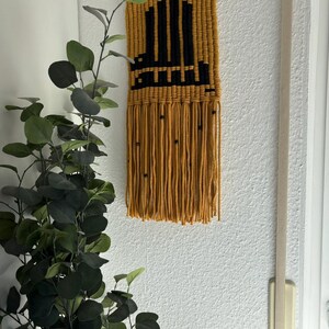 Può includere: Appendiabiti da parete in macramè giallo senape con un design geometrico nero. L'arte murale presenta una forma rettangolare con frange nella parte inferiore e piccole perle nere. Un tassello di legno viene utilizzato per appendere.