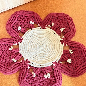 Könnte beinhalten: Handgefertigter, blumenförmiger Teppich in Burgunderrot und Creme. Der Teppich hat einen cremefarbenen Mittelkreis, umgeben von burgunderroten Blütenblättern mit weißen Akzenten. Ein dekoratives Element für Wohnräume.