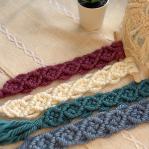 May include: Four macrame wall hangings in different colors: burgundy, cream, teal, and light blue. Each hanging features a repeating pattern of knots.
